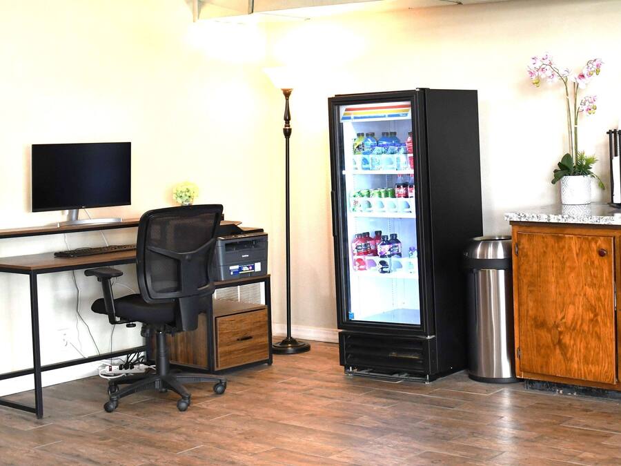 A room with a desk, chair, computer, refrigerator, trash can, and a cabinet.