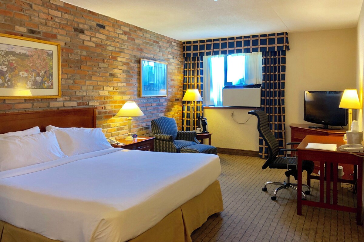 A hotel room with a large bed, two chairs, a desk with a television, and a brick wall.