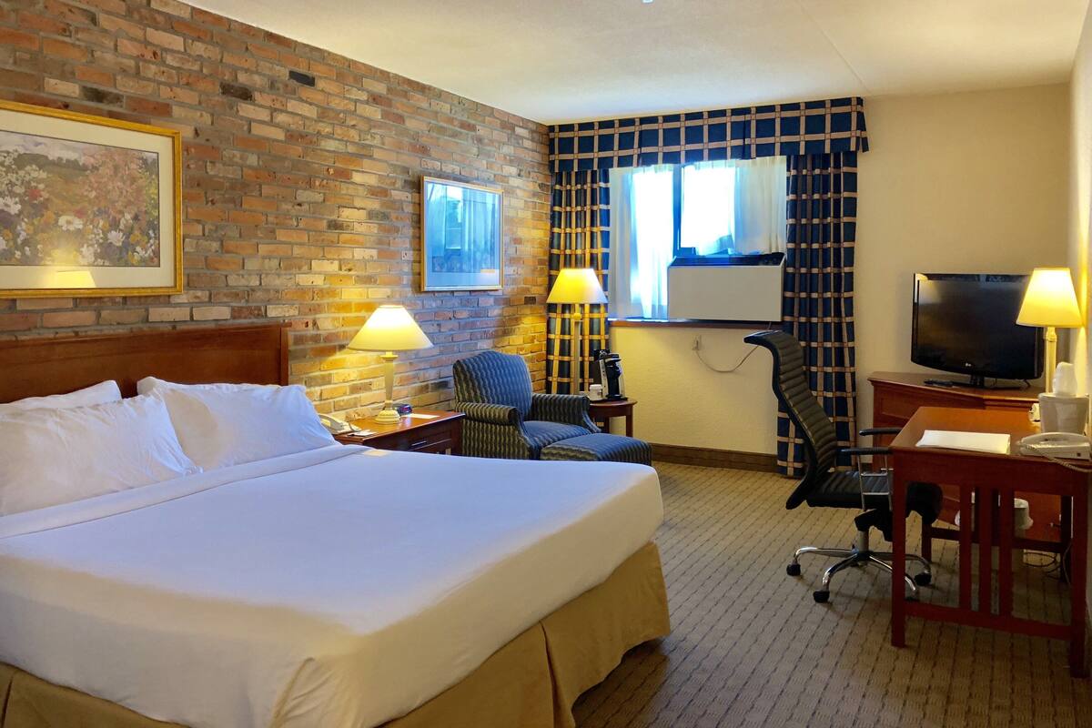 A hotel room with a large bed, two chairs, a desk with a television, and a brick wall.