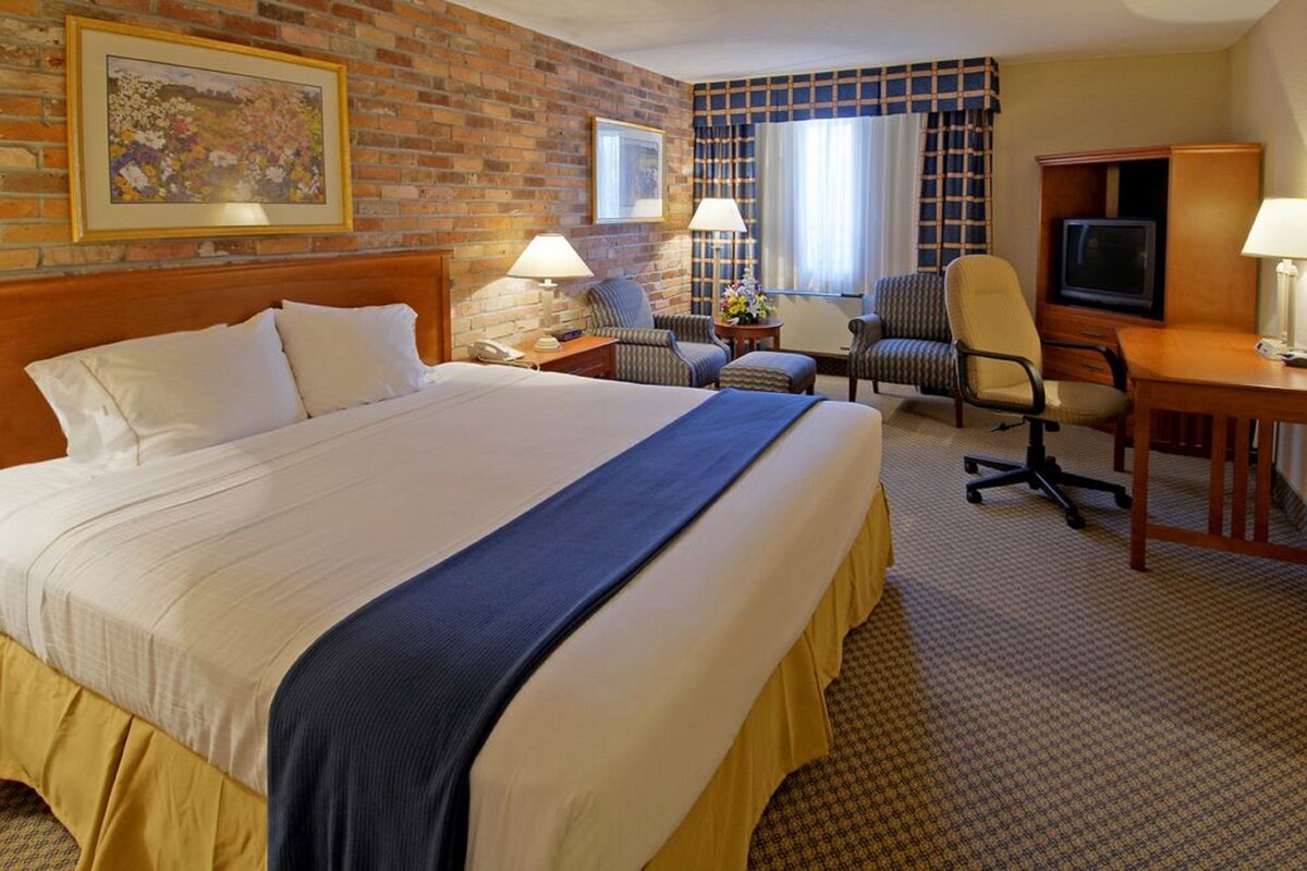 A hotel room with a large bed, two armchairs, a desk with a television, and a brick wall with a framed picture.