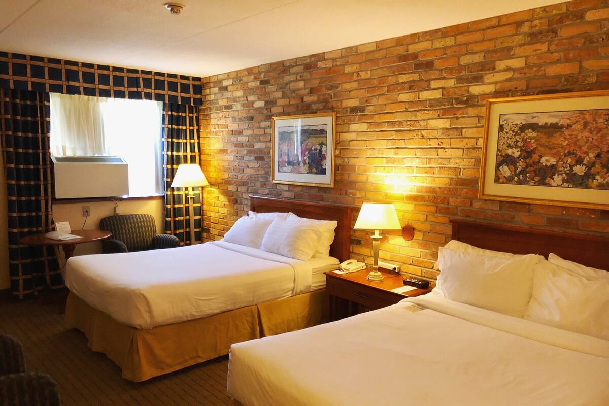 A hotel room with two beds, a brick wall, a window with curtains, a small table, a chair, and a lamp.
