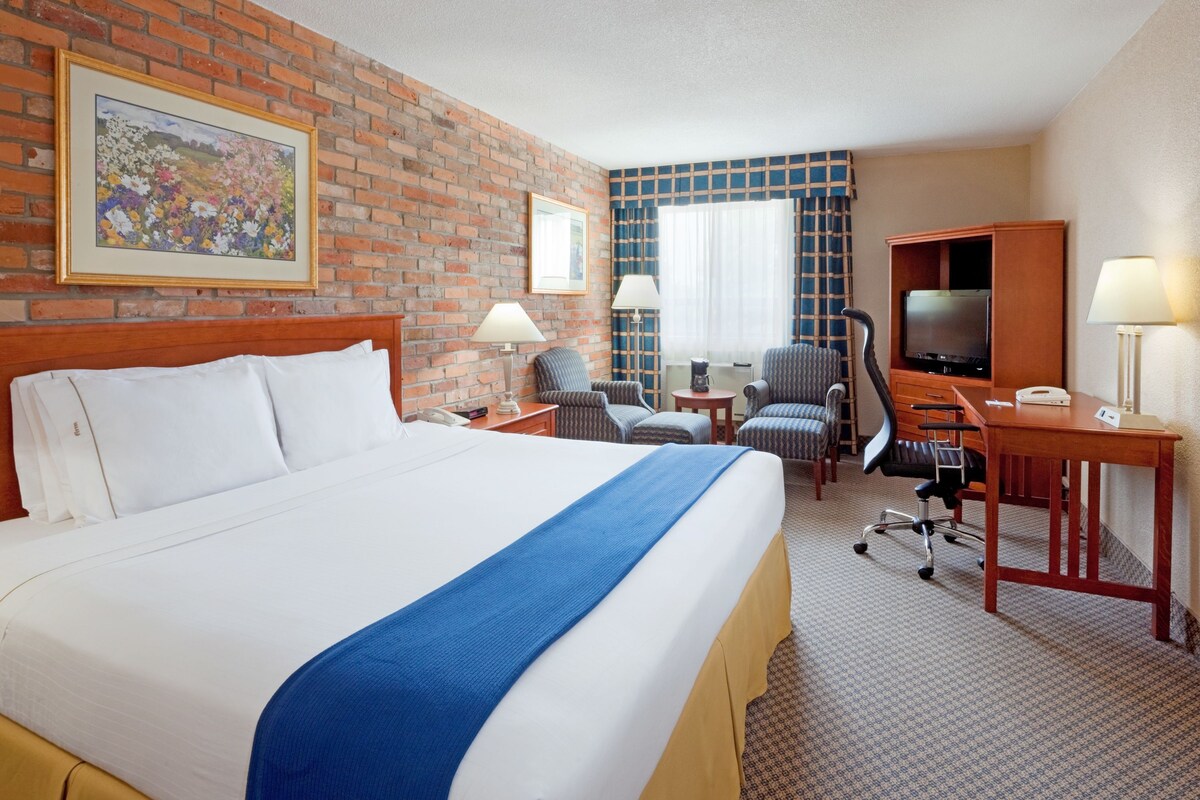 A hotel room with a large bed, a desk with a computer, a TV, and a seating area with chairs.