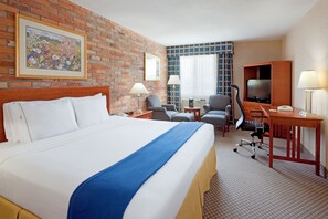 Une chambre d’hôtel avec un grand lit, un bureau avec un ordinateur, une télévision et un coin salon avec des fauteuils.