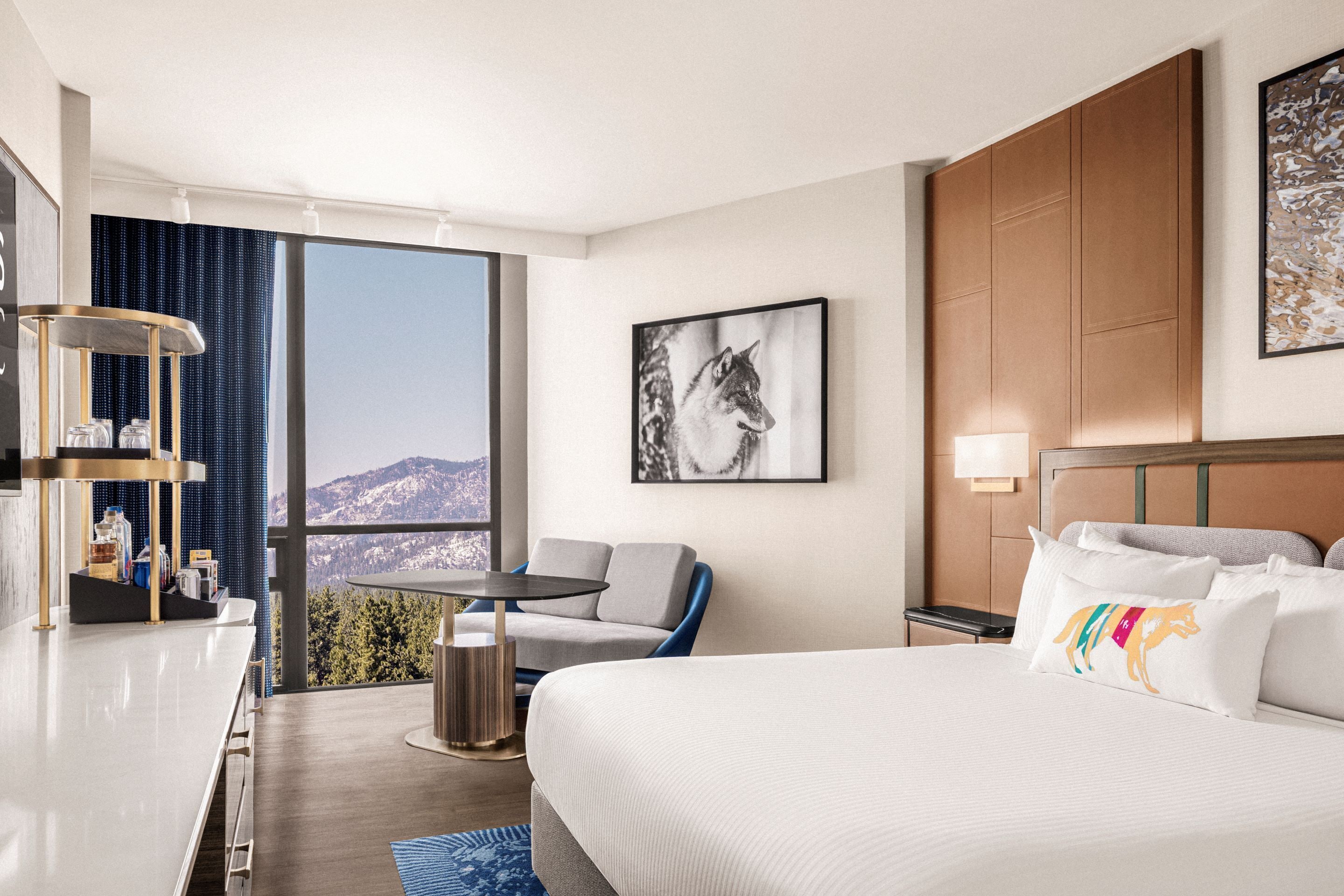 A hotel room with a bed, a desk, a chair, a small table, and a large window offering a mountain view.