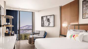 A hotel room with a bed, a desk, a chair, a small table, and a large window offering a mountain view.