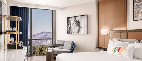 A hotel room with a bed, a desk, a chair, a small table, and a large window offering a mountain view.