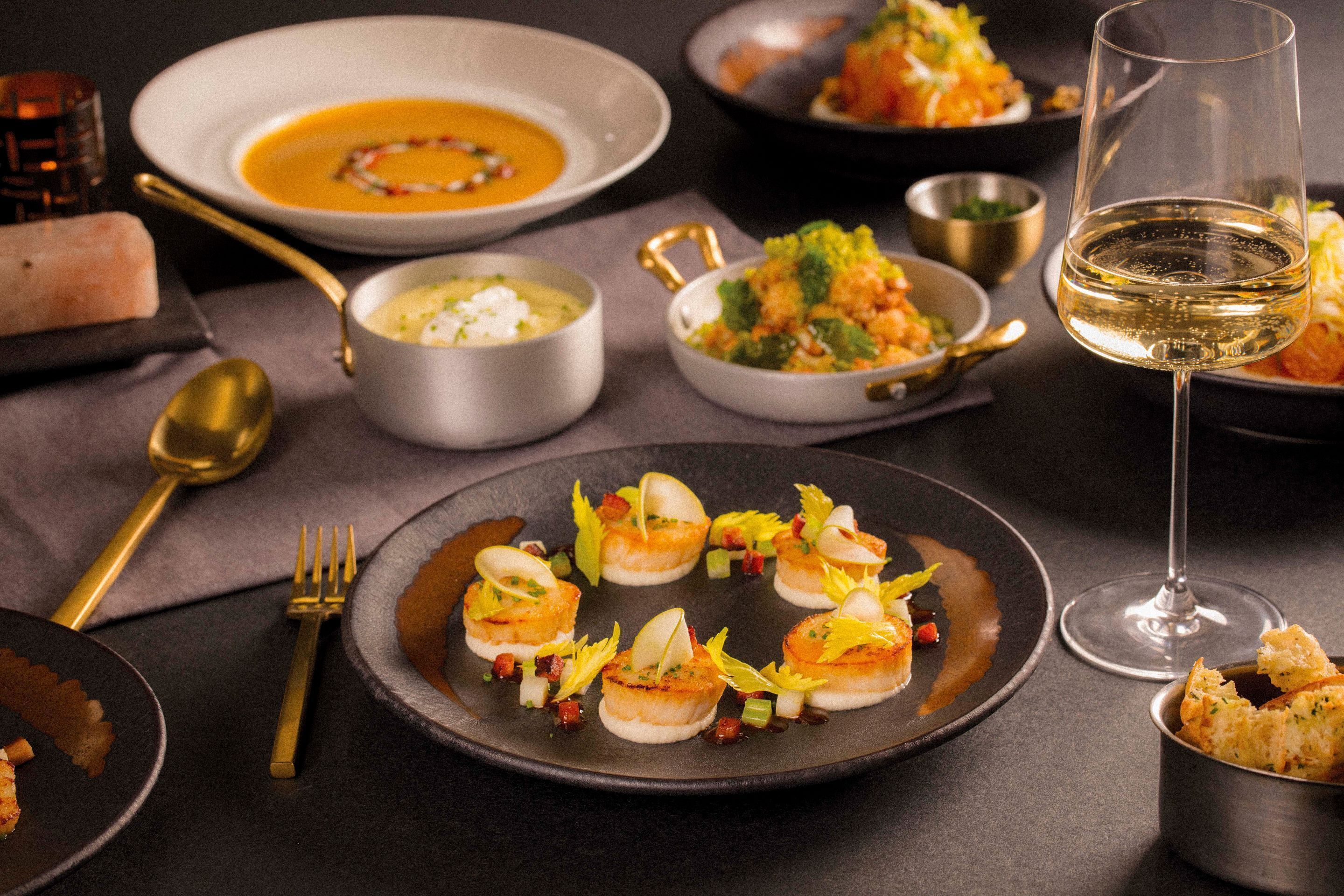 A dining table with various dishes, including a bowl of soup, a plate of scallops, and a glass of wine.