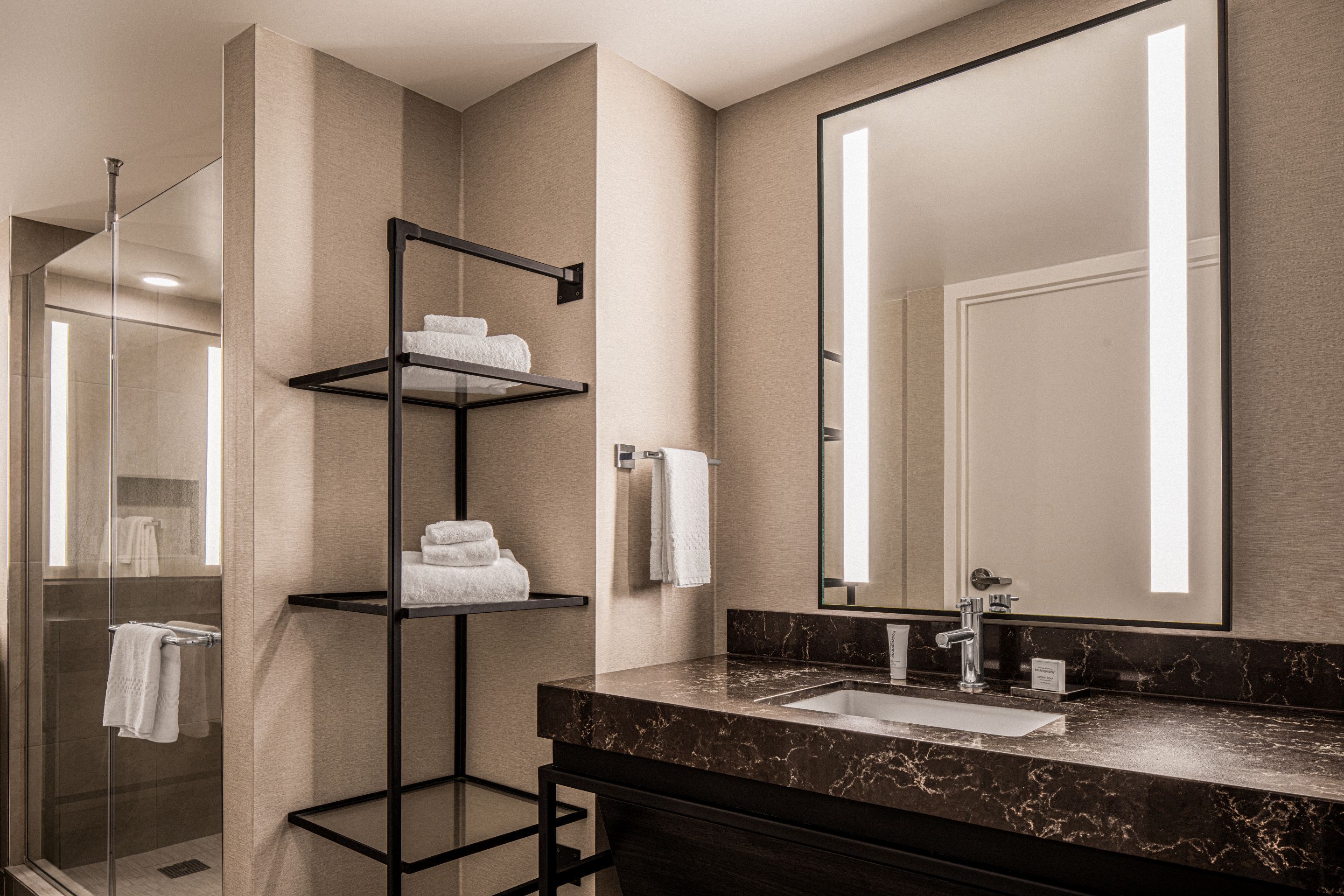 A modern bathroom with a glass shower enclosure, a black metal towel rack, a marble countertop, and a large mirror.