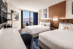 Habitación de hotel con dos camas, un escritorio, una silla, un televisor y un ventanal con vistas a las montañas.