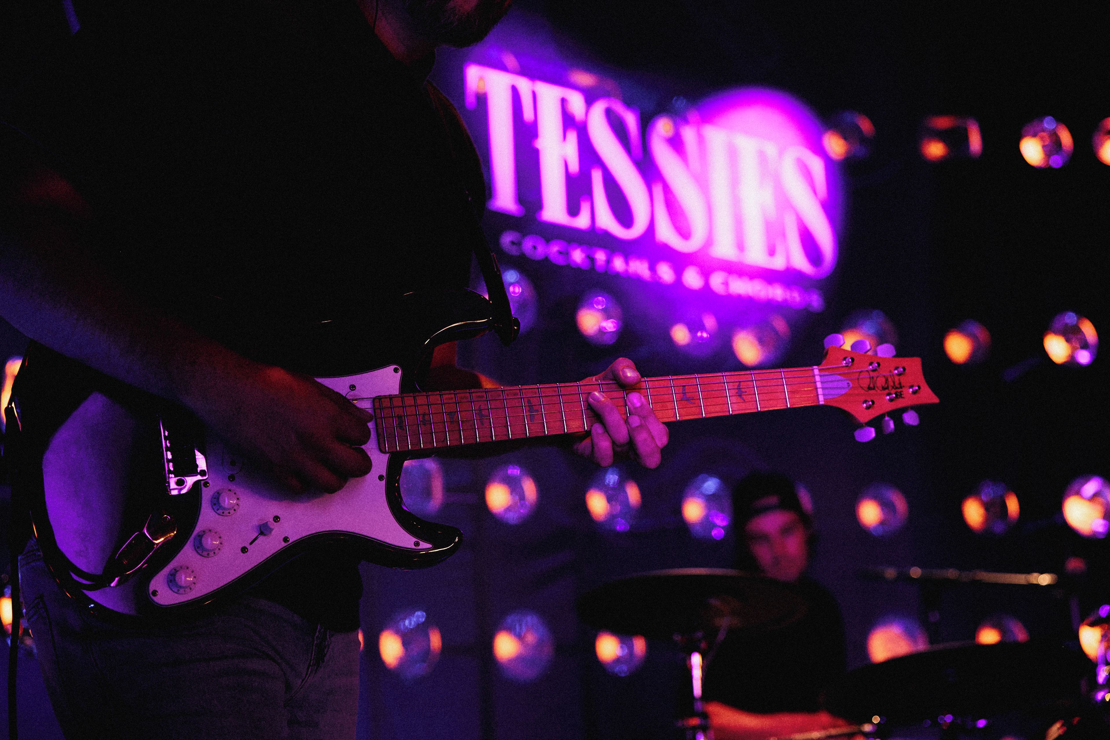 Une personne joue de la guitare électrique sur scène, et on peut voir en arrière-plan une enseigne sur laquelle est écrit : « TESSIE'S COCKTAILS & CIGARS ».