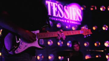 Une personne joue de la guitare électrique sur scène, et on peut voir en arrière-plan une enseigne sur laquelle est écrit : « TESSIE'S COCKTAILS & CIGARS ».