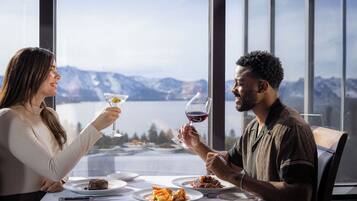 A dining table with plates of food and wine glasses, set against a backdrop of mountains and a lake.