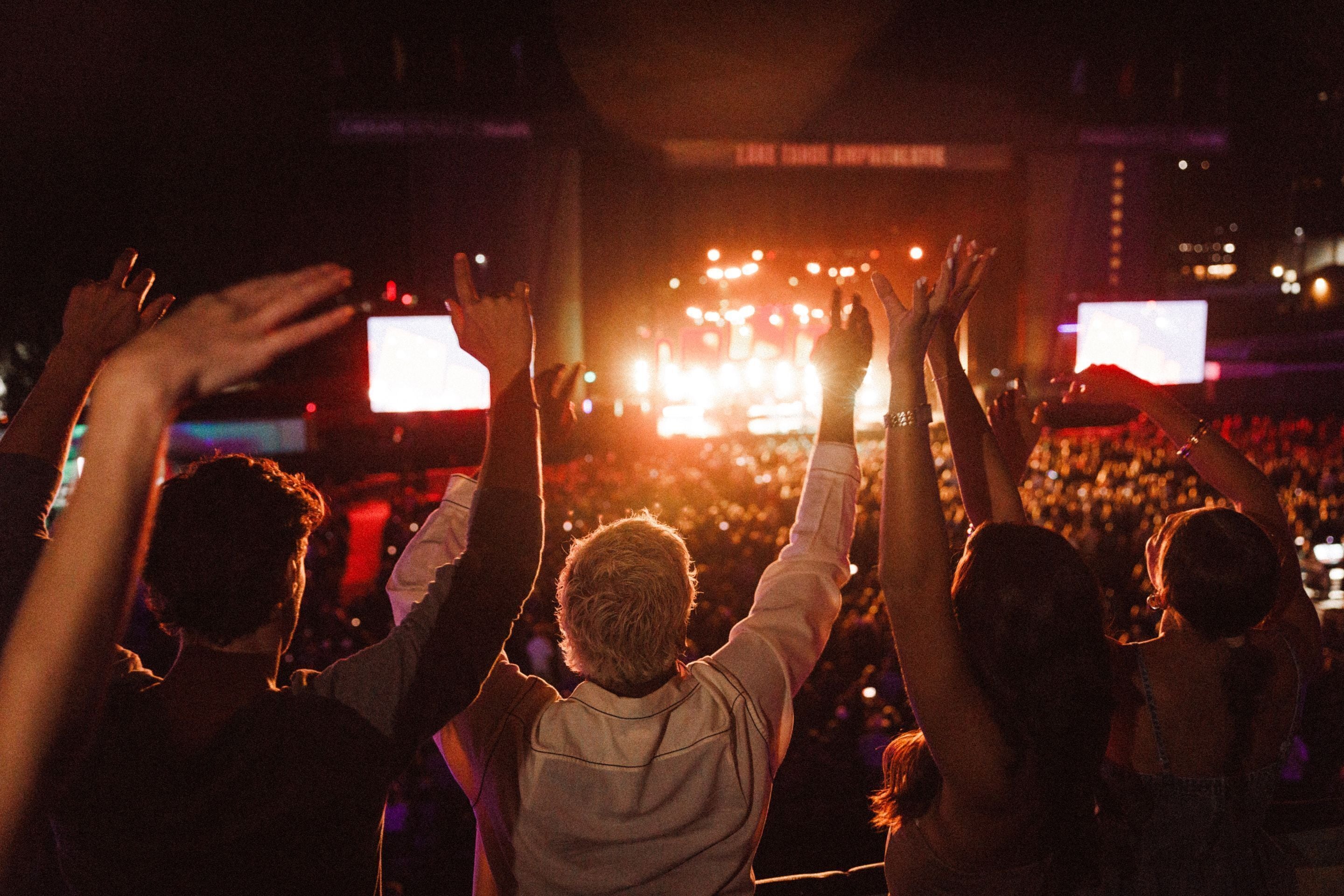 A crowd at a concert with raised hands, a stage with bright lights, and a large screen displaying visuals.