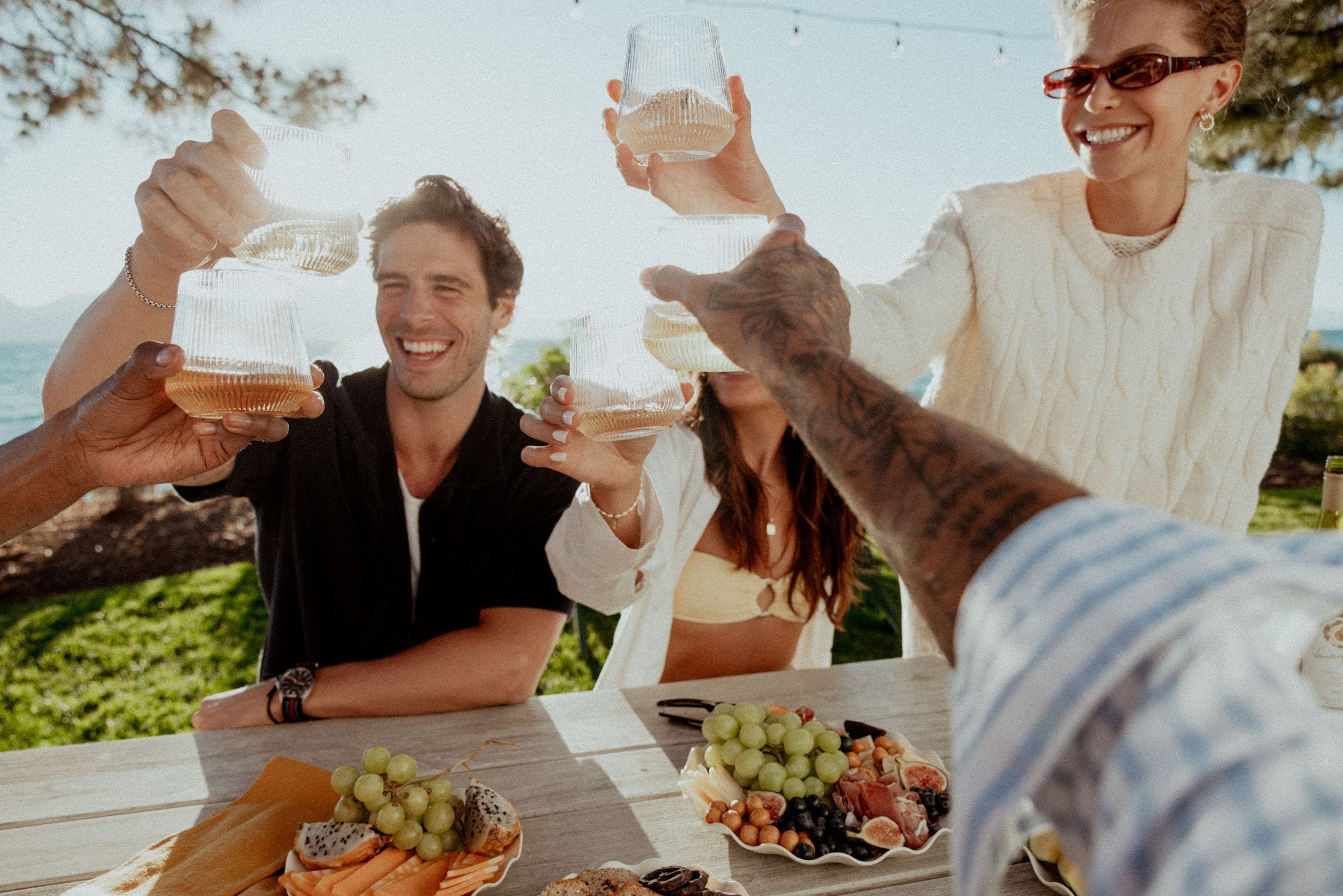 A group of people toasting with glasses at an outdoor table with a variety of fruits and cheeses.