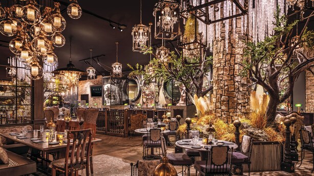 A restaurant with multiple dining tables, chairs, hanging chandeliers, and a stone wall backdrop.