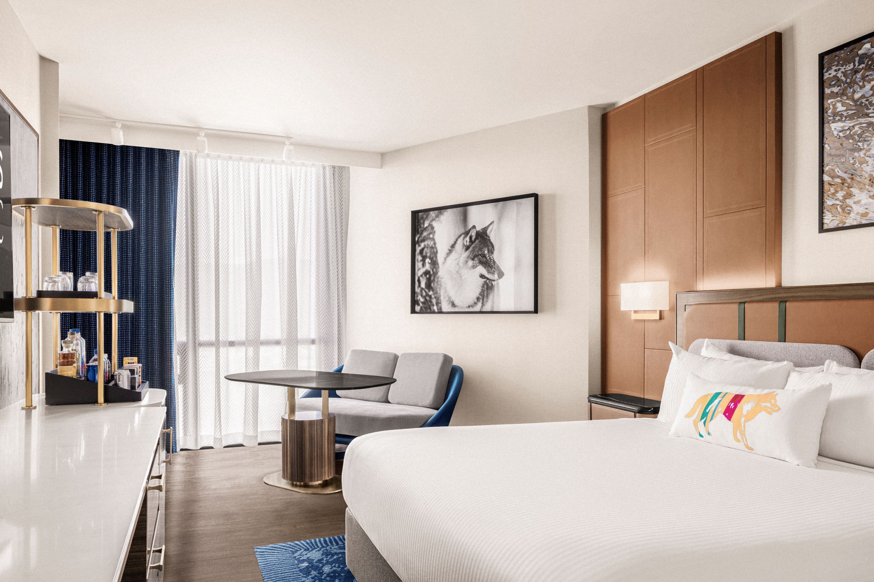 Una habitación de hotel moderna con una cama grande, un escritorio, una silla y una mesita pequeña. Hay una ventana con cortinas, una lámpara de pared e imagen enmarcada en la pared.