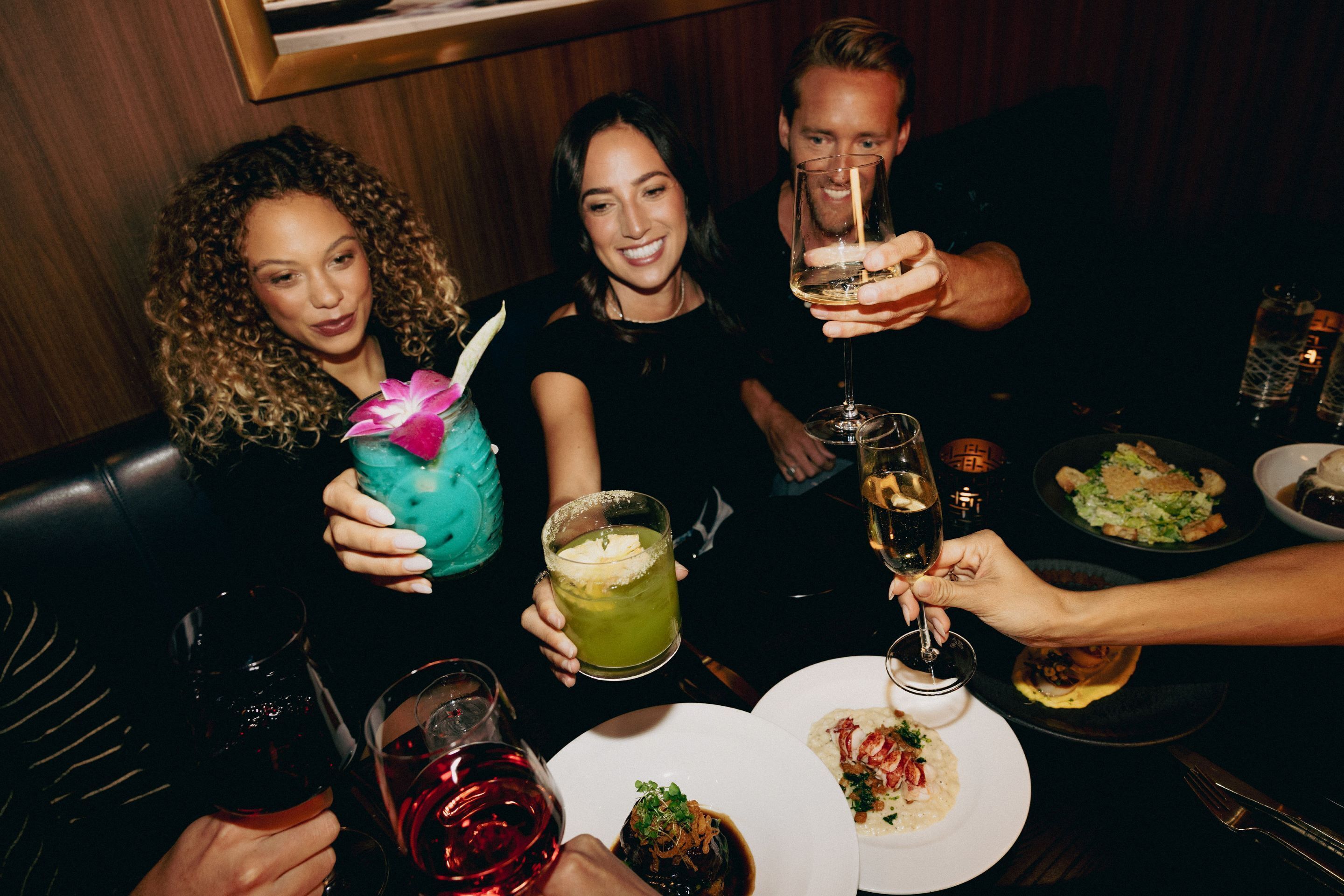 A group of people seated at a table with drinks and food, in a dimly lit indoor setting.