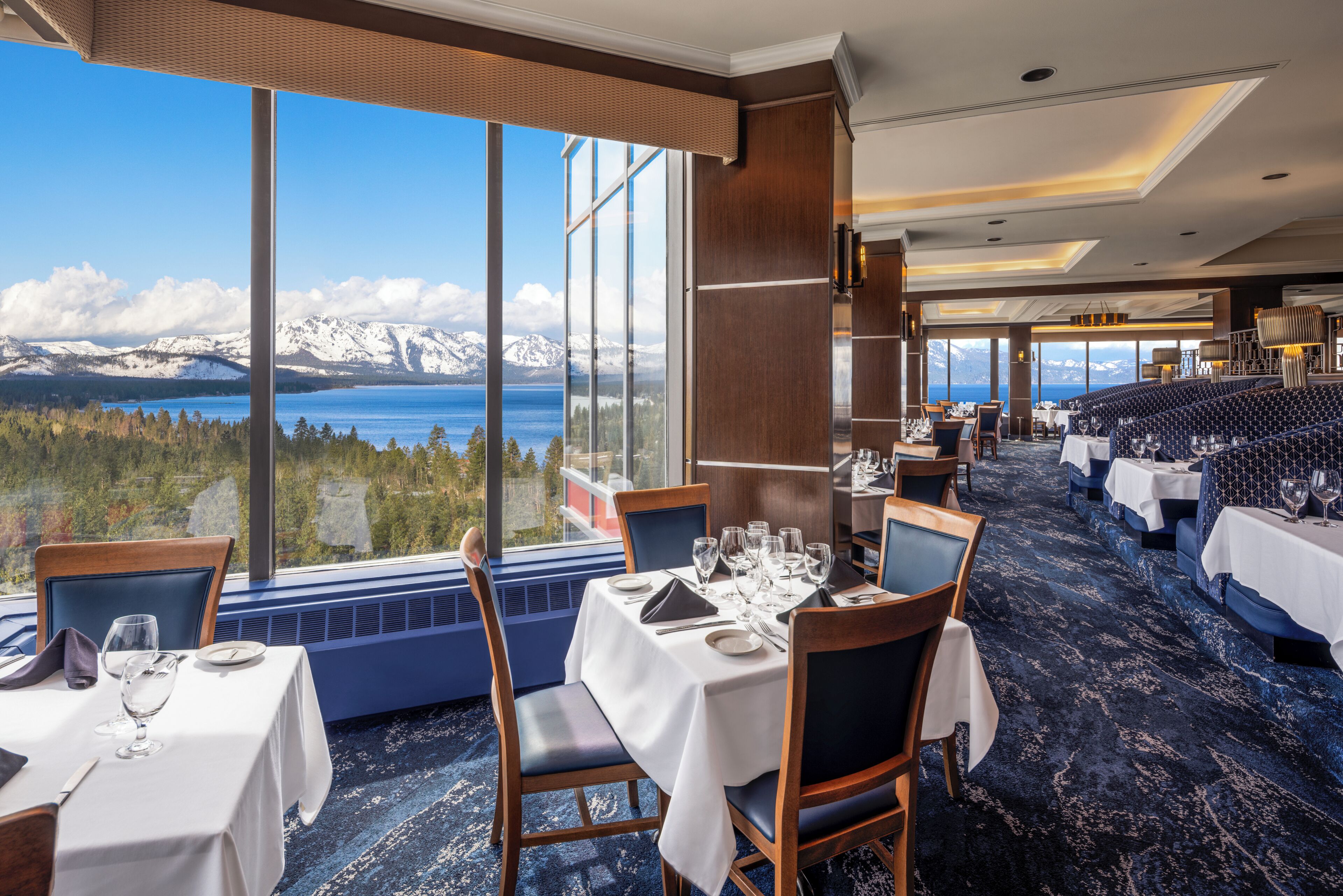A restaurant with a view of mountains and a lake, tables set with white tablecloths, and blue chairs.
