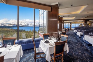 A restaurant with a view of mountains and a lake, tables set with white tablecloths, and blue chairs.