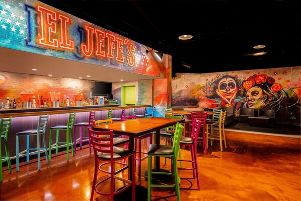 Un área de bar vibrante con un letrero de neón que dice "EL JEFE'S" y sillas y mesas coloridas.