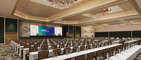 Un salón de conferencias con filas de mesas y sillas, una pantalla grande mostrando una presentación y un candelabro.