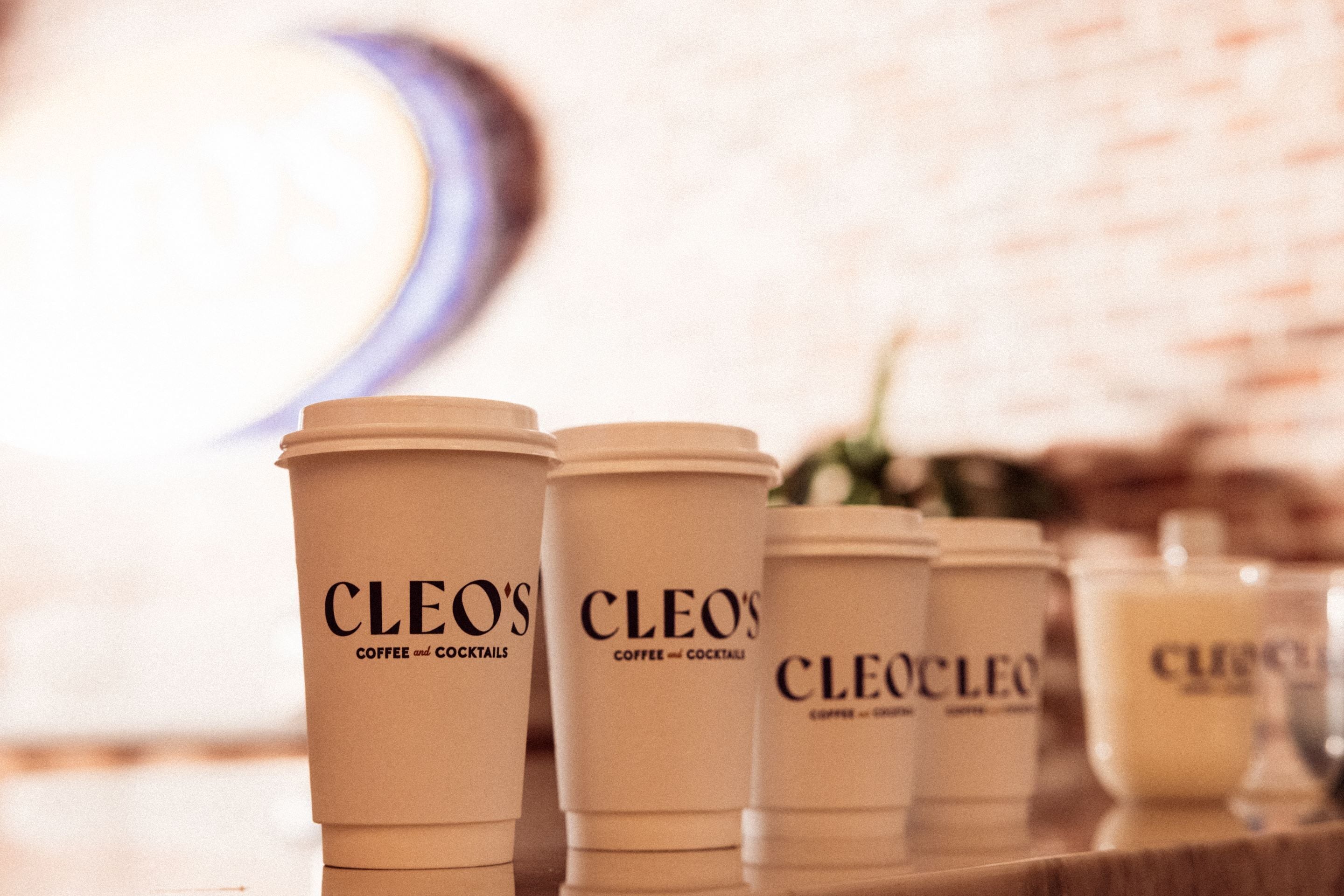 A row of coffee cups labeled "CLEO'S COFFEE and COCKTAILS" on a counter.