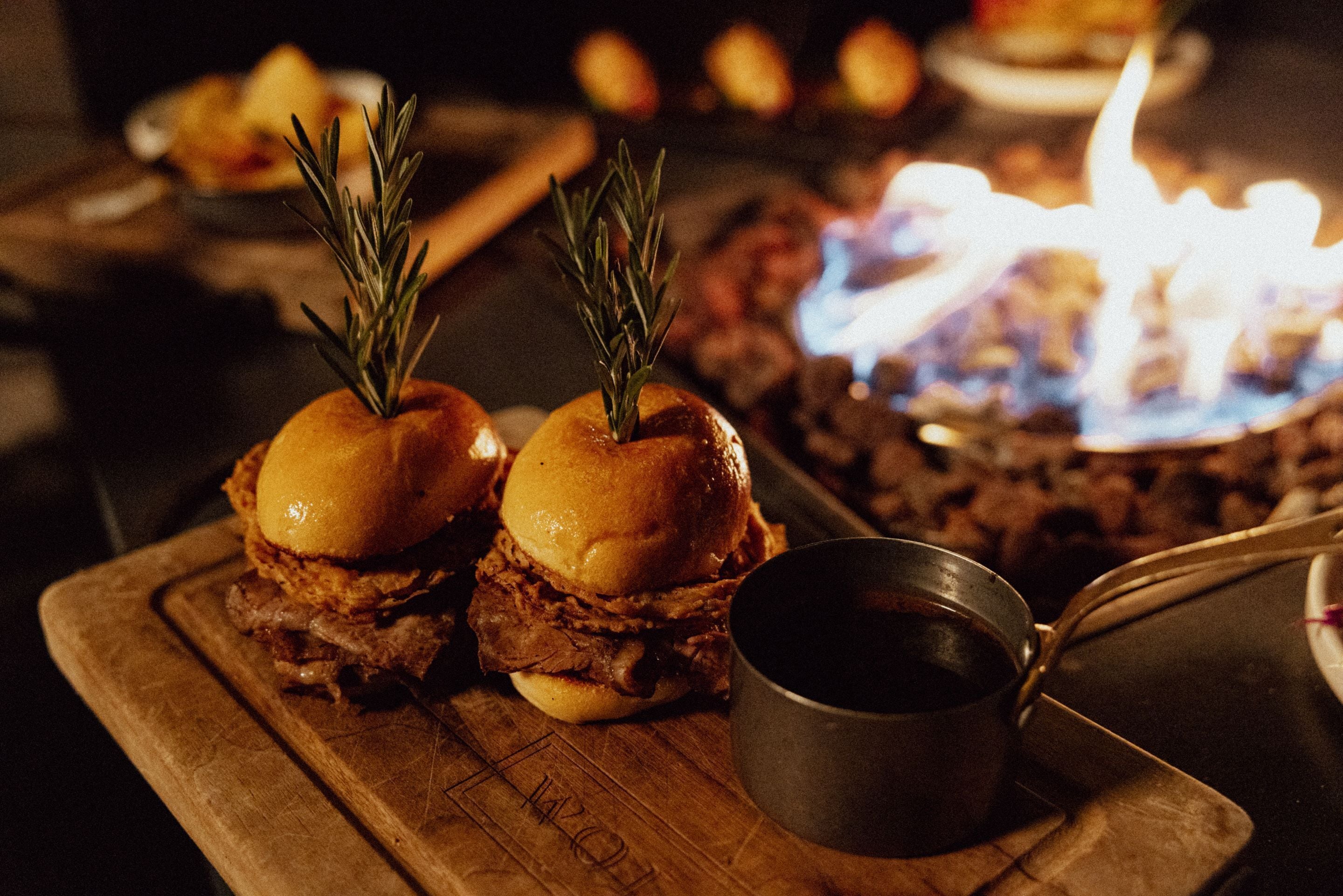 Two sandwiches with rosemary on a wooden board, a metal cup, and a lit fire in the background.