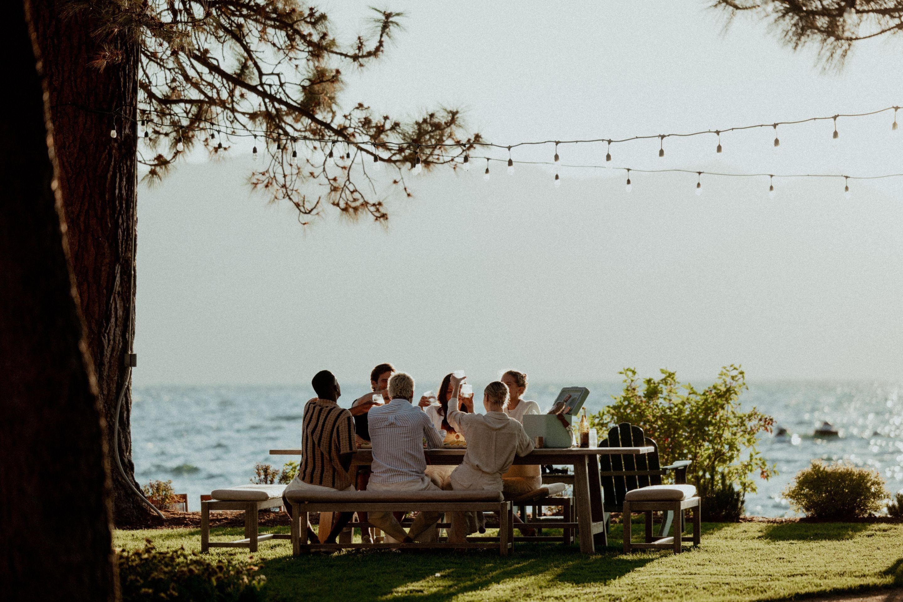 A group of people seated at a wooden table outdoors with a view of the ocean.