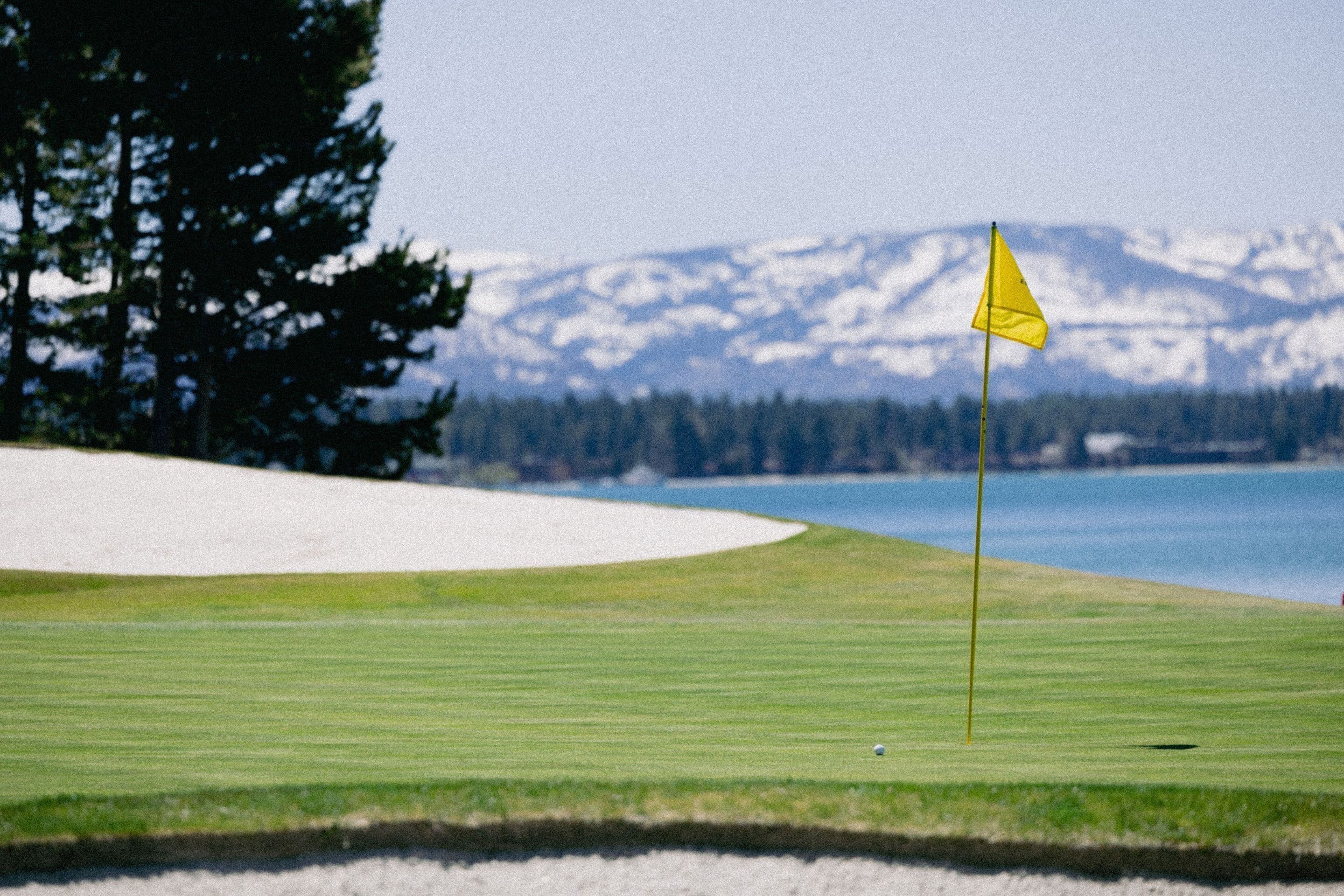 Un campo de golf con un mástil de bandera, un búnker de arena y montañas al fondo.