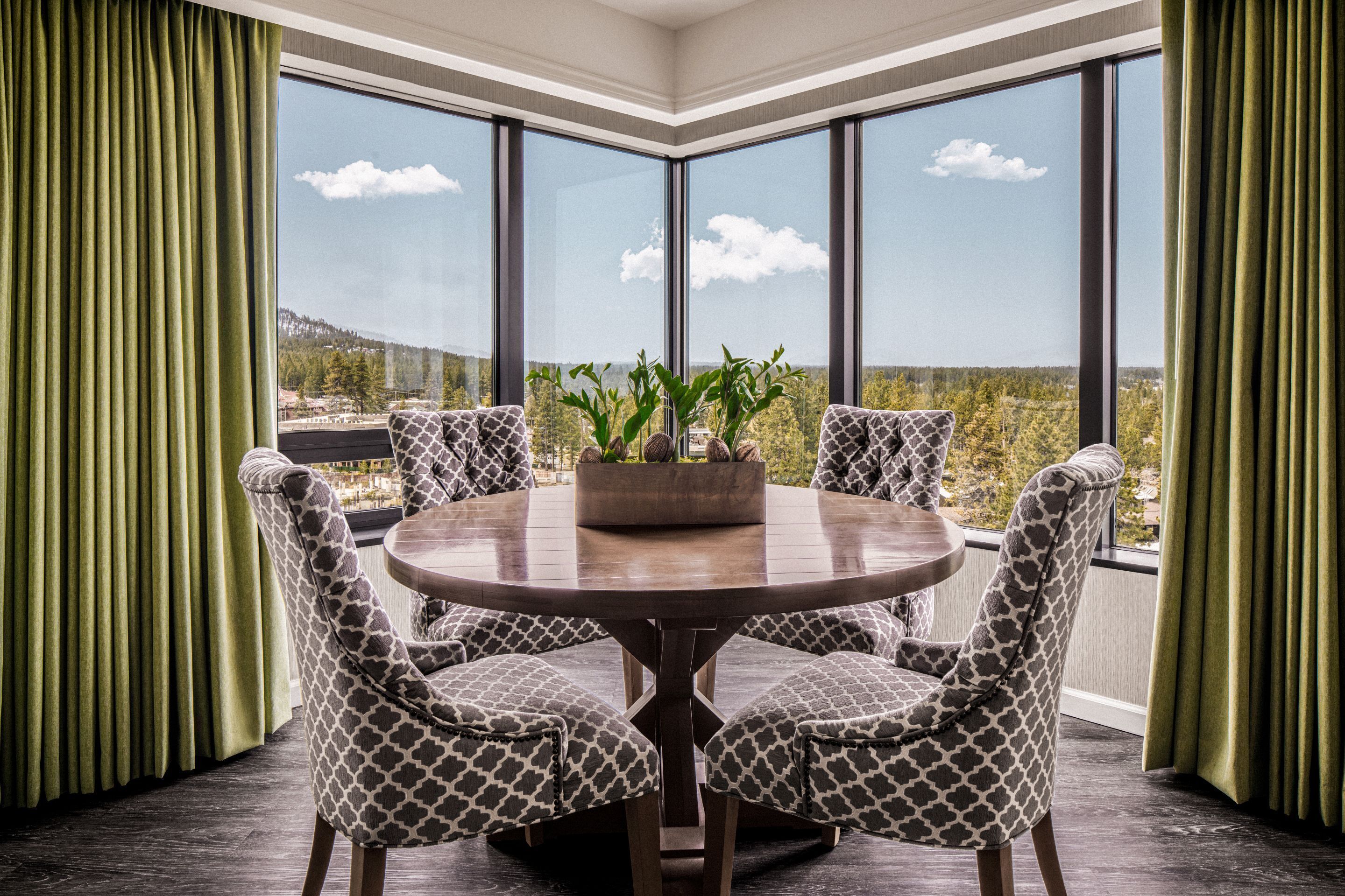 A corner room with a round table, four patterned chairs, a potted plant, and a view of a mountainous landscape through large windows.