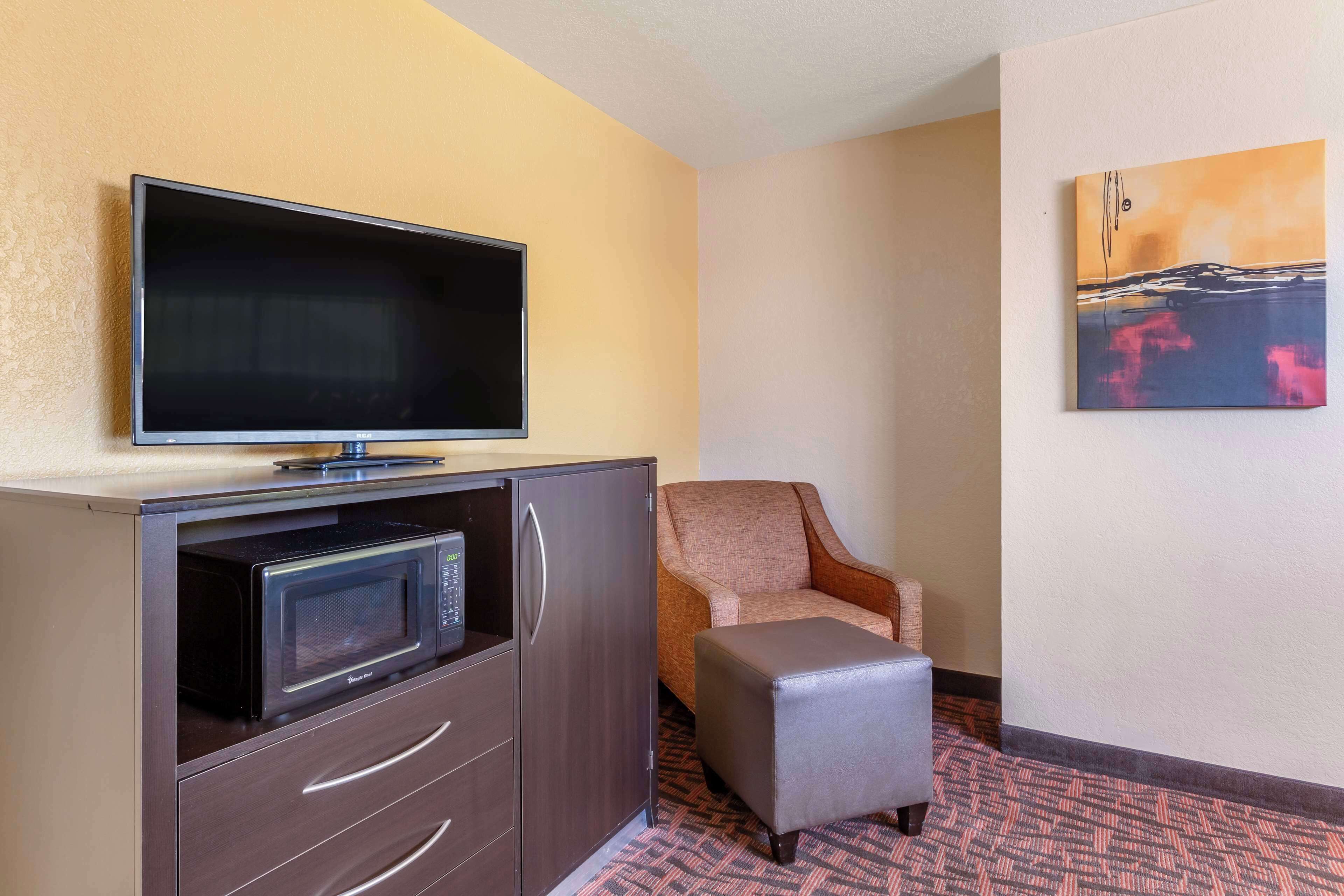A hotel room with a flat-screen TV on a cabinet, a microwave, a chair, an ottoman, and a painting on the wall.