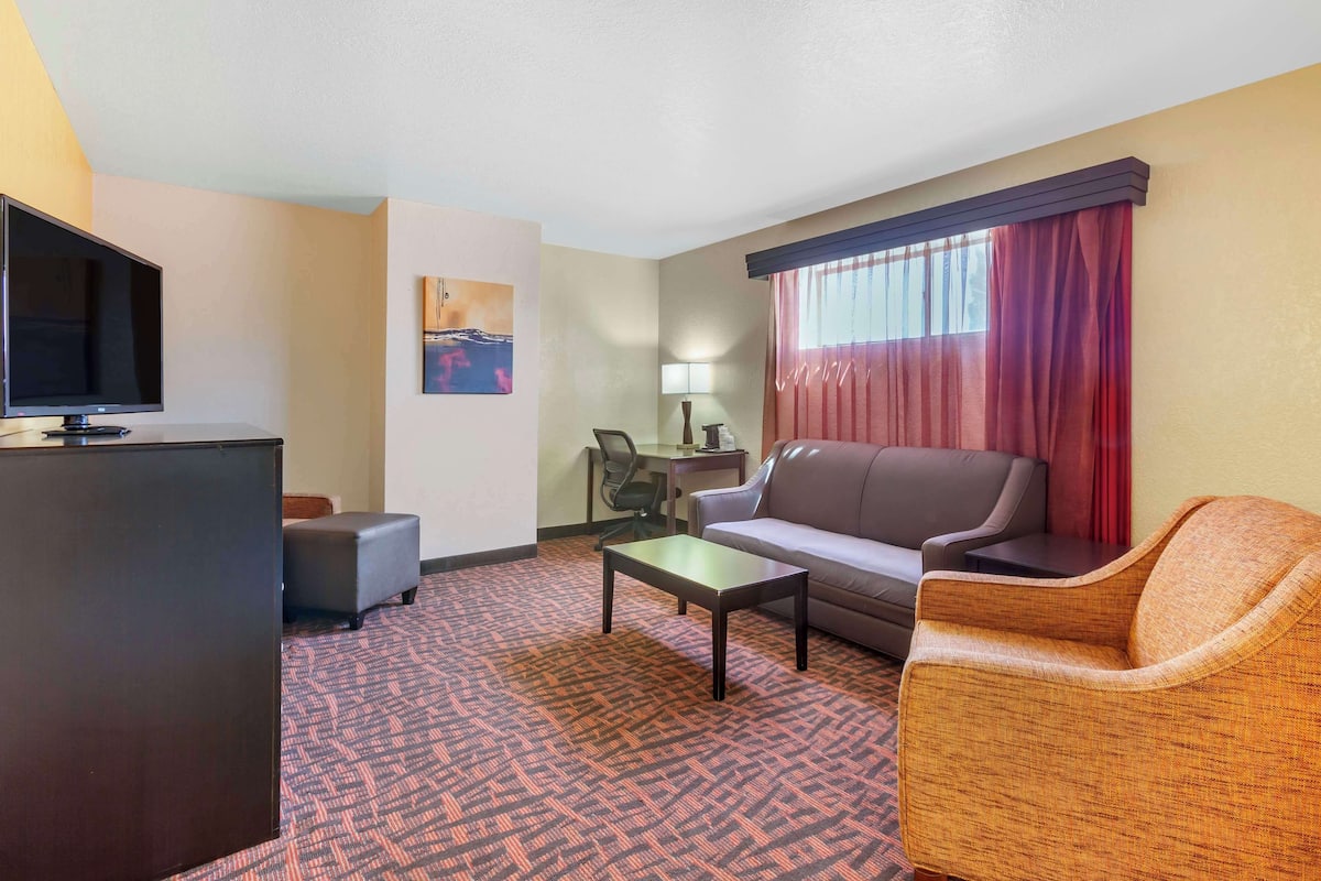 A hotel room with a flat-screen TV, a sofa, a loveseat, a small table, a desk with a chair, and a patterned carpet.