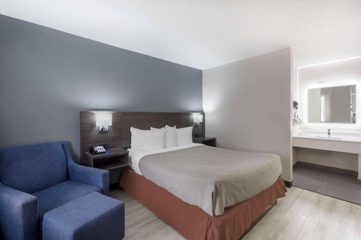 A hotel room with a bed, bedside table, lamp, chair, and a bathroom with a mirror and sink.