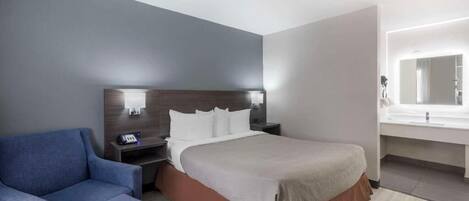 A hotel room with a bed, bedside table, lamp, chair, and a bathroom with a mirror and sink.