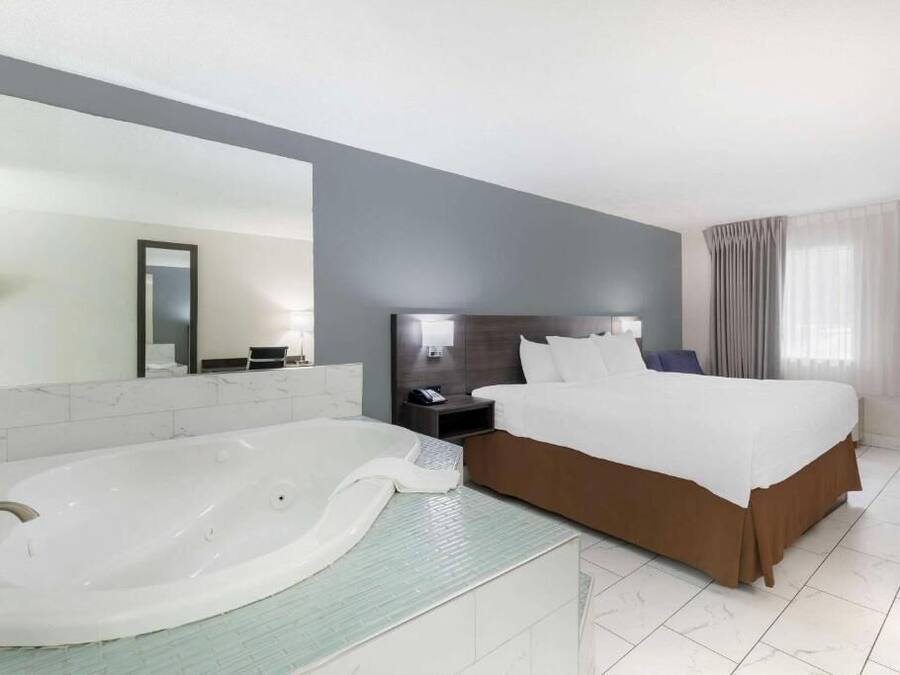 A modern hotel room with a large bed, a separate bathroom with a bathtub, a large mirror, and a nightstand with a lamp.