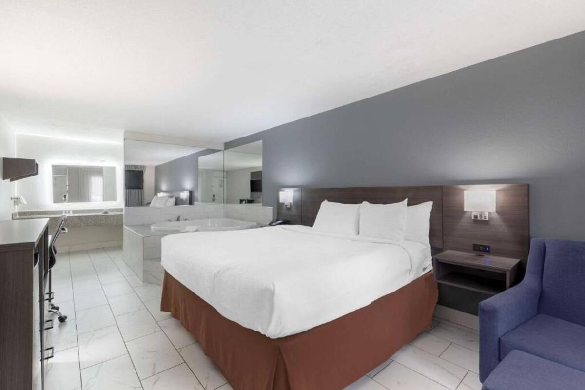 A modern hotel room with a large bed, a bedside table, a chair, a desk, and a bathroom with a mirror and sink.