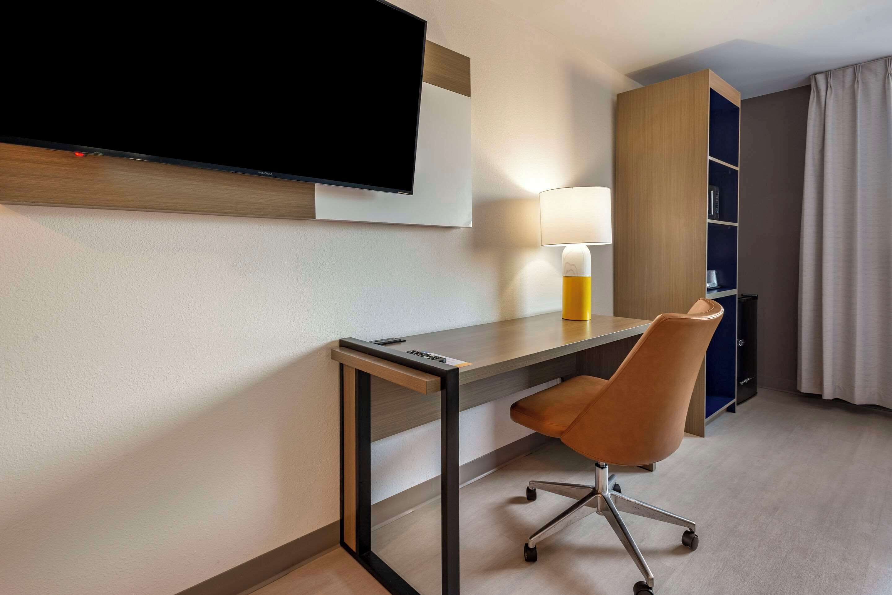 A hotel room with a flat-screen TV mounted on the wall, a desk with a chair, a lamp, and a bookshelf.
