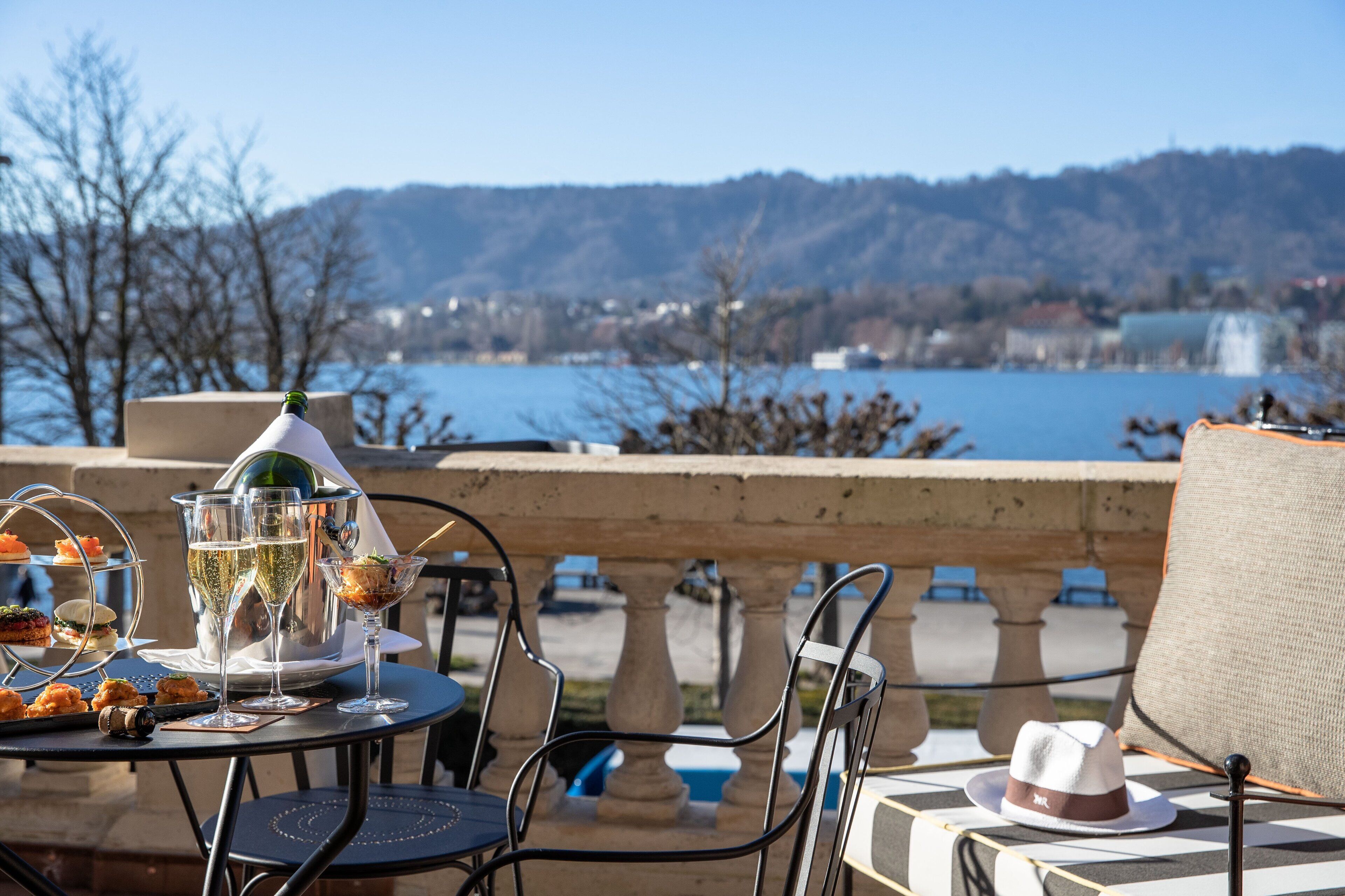 Une table dressée avec une bouteille, des verres et un plateau de nourriture, donnant sur un lac et des collines.