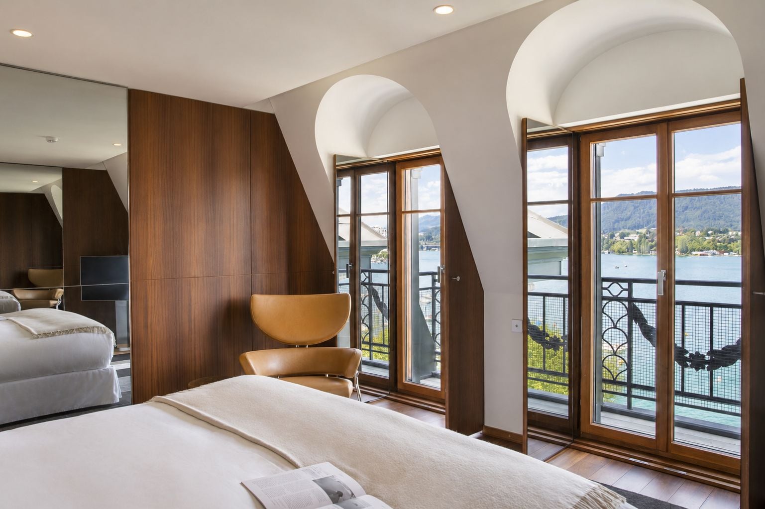 A modern hotel room with a large bed, a seating area, and a balcony with a view of a lake.