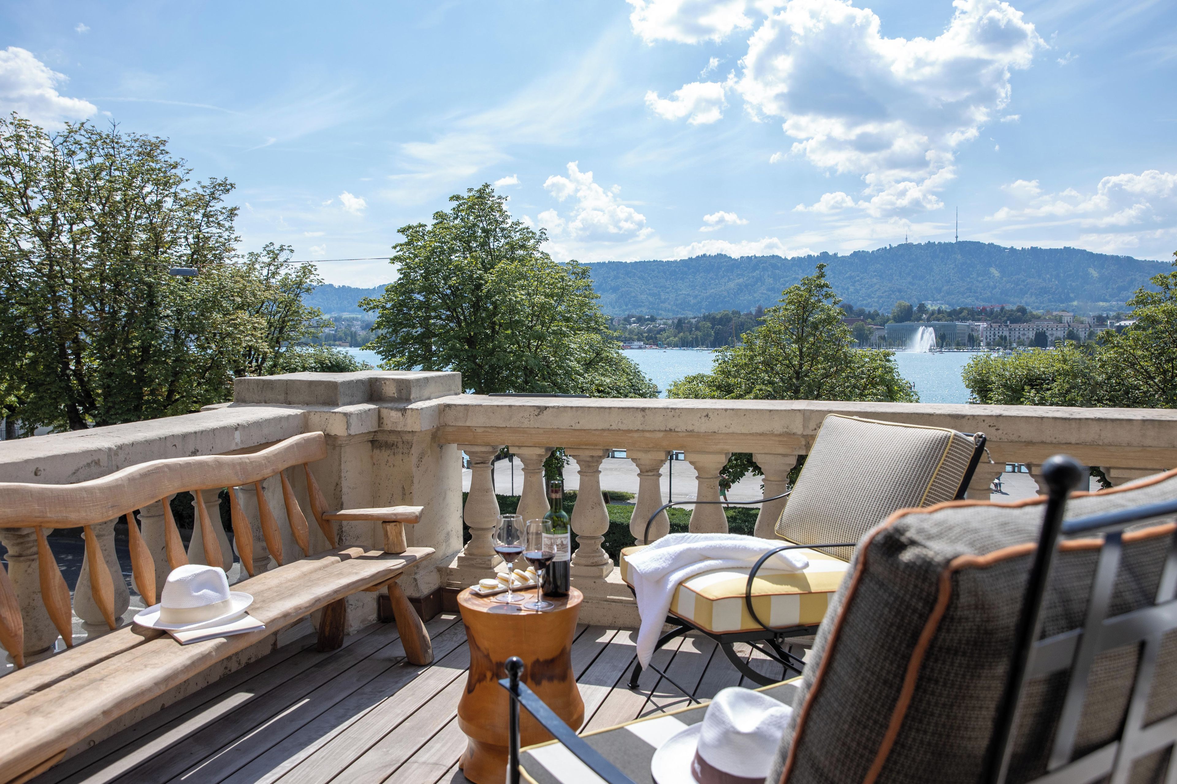 Eine Terrasse mit Blick auf einen See und Berge, ausgestattet mit einer Bank, Stühlen und einem Tisch mit Weingläsern und einer Weinflasche.