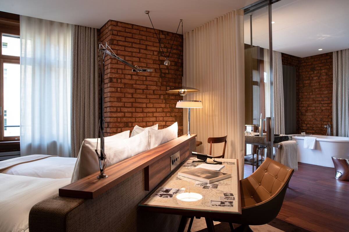 Een hotelkamer met een bakstenen muur, een bed met witte lakens, een houten bankje, een tafel met een lamp en een stoel.
