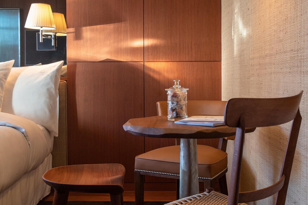 Una habitación de hotel con una cama, una silla de madera, una mesita redonda con una lámpara y un aplique de pared.