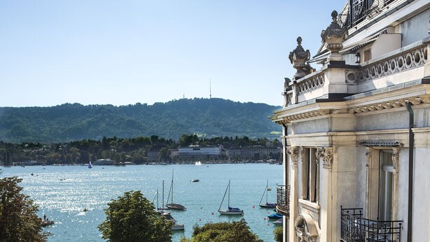 A coastal view with boats on the water, a building with ornate balconies, and a hill in the background.