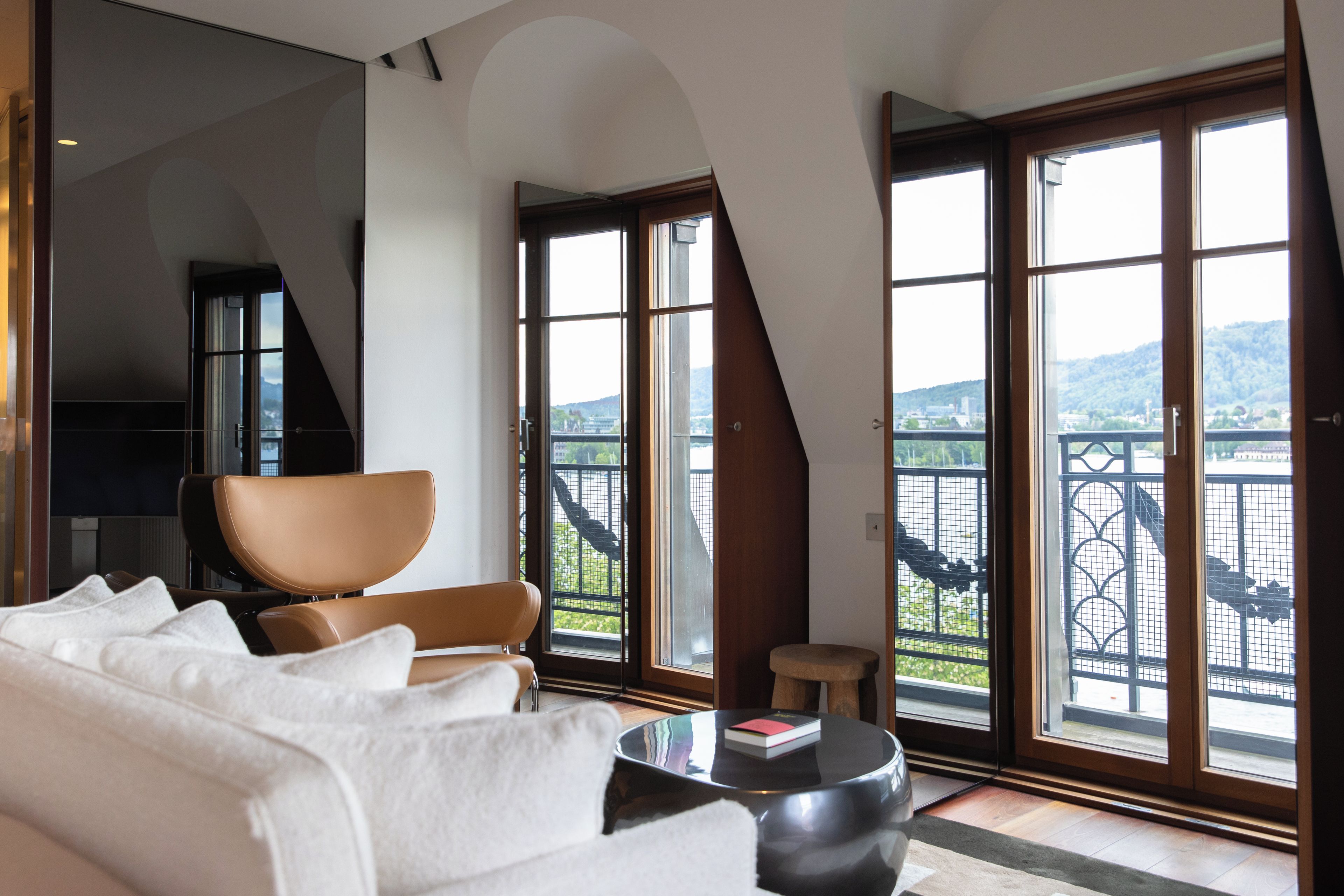 A modern living room with a sofa, a chair, a small table with a book, and large windows with a view of a balcony and landscape.