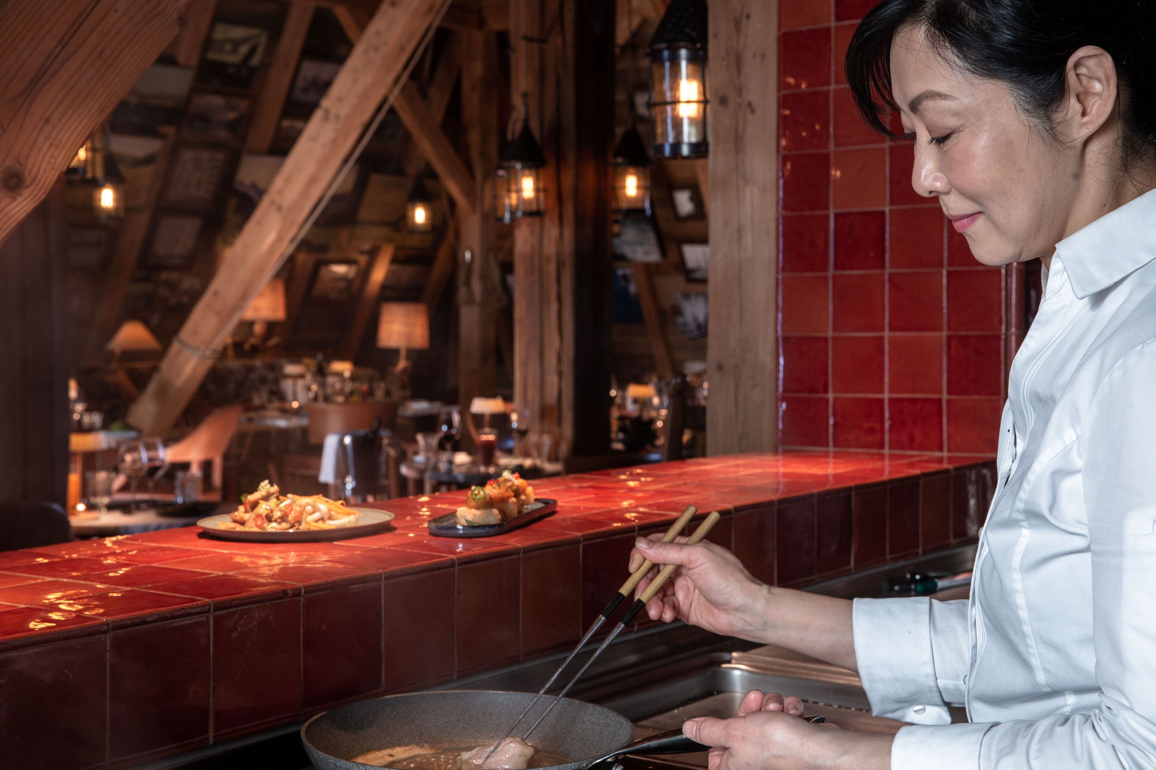 Une personne cuisine dans une cuisine dont le mur derrière l’évier est recouvert de carreaux rouges et qui possède des poutres en bois.