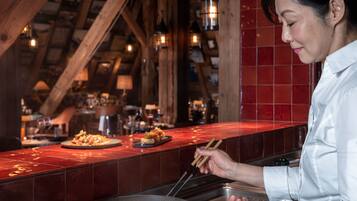 Une personne cuisine dans une cuisine dont le mur derrière l’évier est recouvert de carreaux rouges et qui possède des poutres en bois.