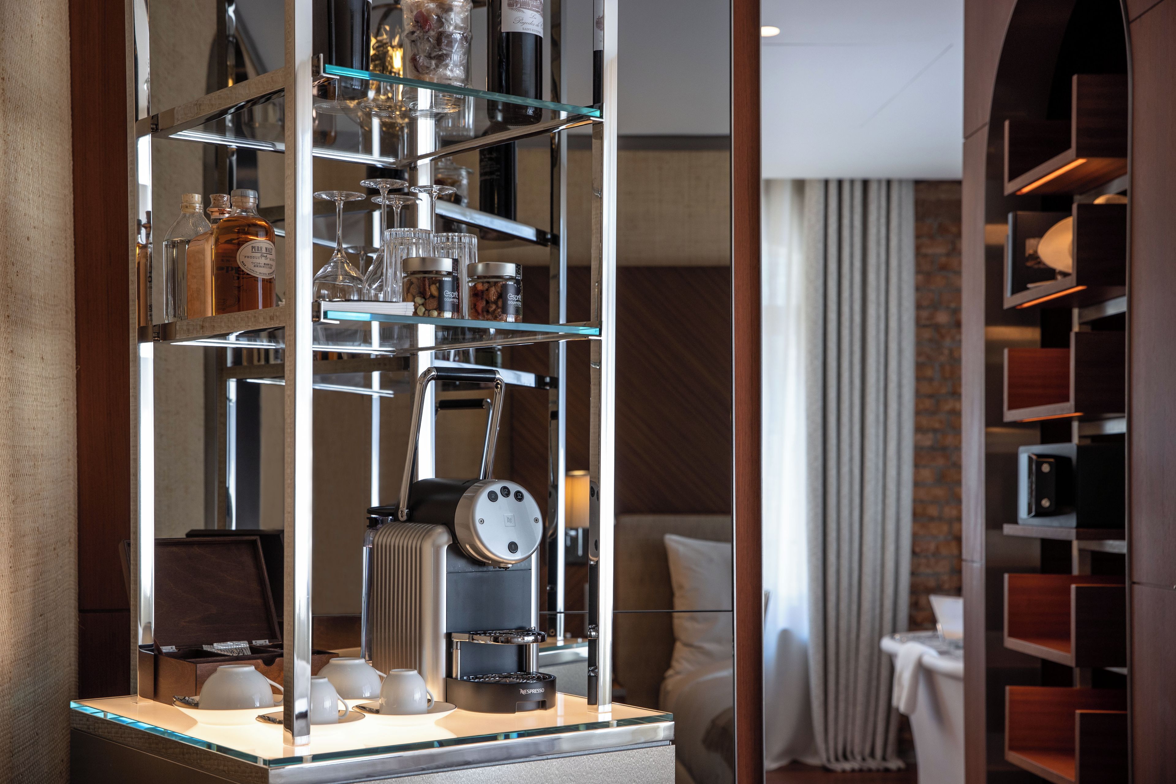 A modern hotel room with a glass display case containing various items, a Nespresso coffee machine, and a cozy seating area with a view of the city.