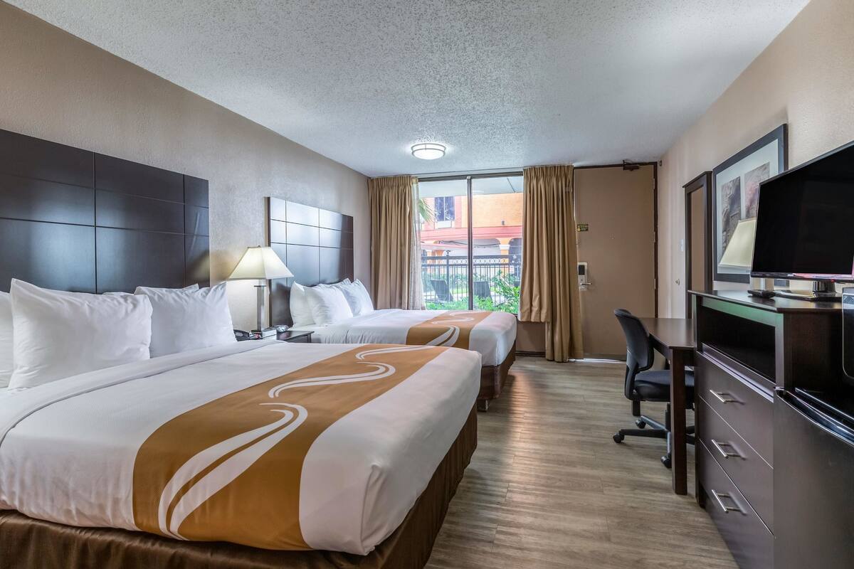 A hotel room with two beds, a desk, a chair, a television, and a large window with curtains.