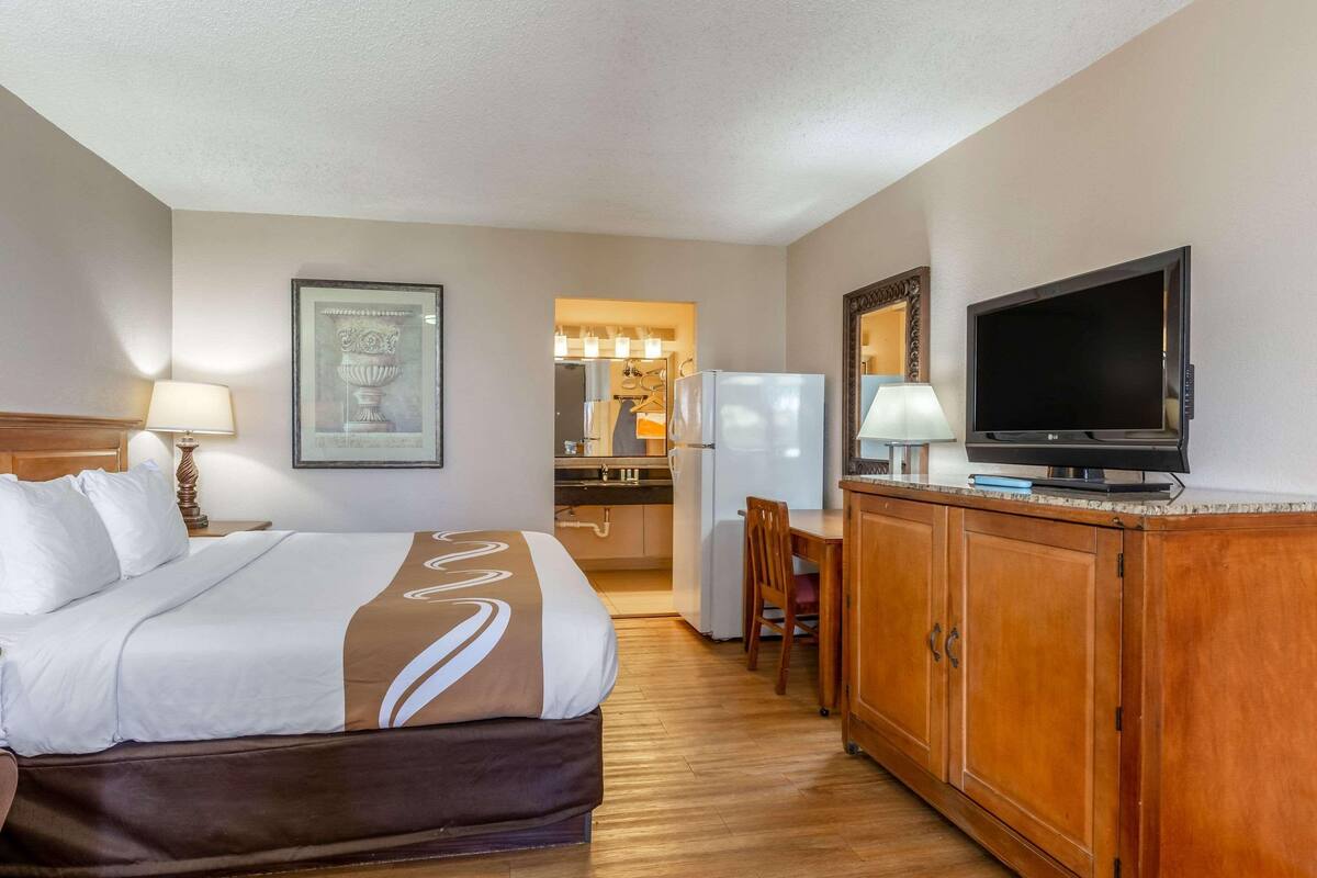 A hotel room with a bed, a television, a desk, a chair, a refrigerator, and a kitchen area.