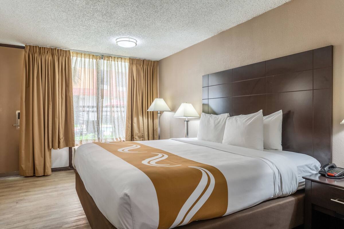 A hotel room with a large bed, two bedside lamps, a headboard, and a window with curtains.